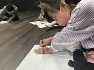 마스크를 쓴 꼬마아이가 종이에 한 손을 대고 다른 손으로 연필을 잡고 종이 위의 손을 따라 그리는 사진